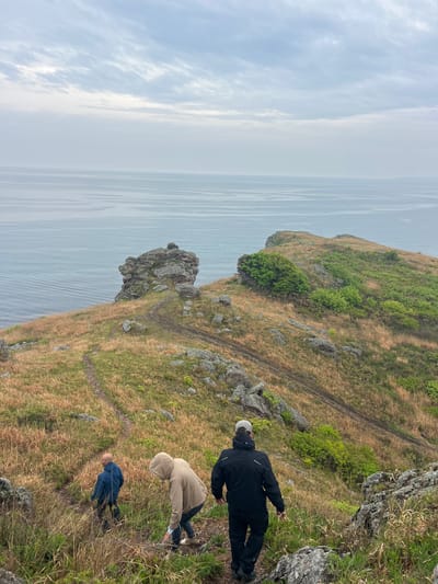 The image shows three people on a grassy cliff overlooking the ocean. A person in a black jacket and light cap is near the edge of the cliff on the right. Below them, another person is wearing a tan jacket with a hood. Further down the slope is a person in a blue jacket. The landscape consists of dry grass, rocks, and patches of green vegetation. The sea is visible in the background, under an overcast sky. The location is Gvozdevo, Russia.