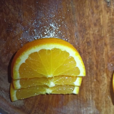 The image shows a close-up, overhead view of three slices of an orange stacked on a wooden surface. The orange slices are a bright yellow-orange color, and the rind is visible on the top slice. The wooden surface appears to be a cutting board with visible grain and some water droplets. The lighting creates small, blurry highlights on the orange and the wood. There are no people or any other objects visible in the image. The scene is indoors, likely in a kitchen. The location is not identifiable as Limerick, Ireland.