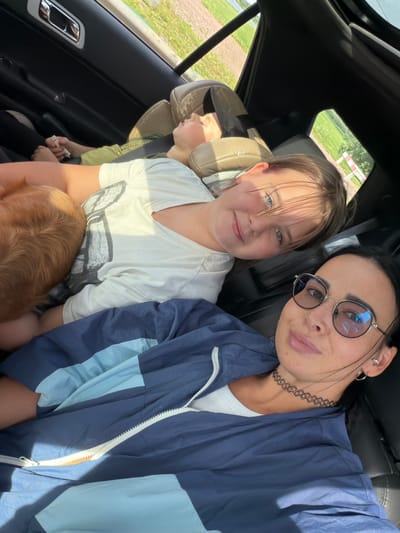 In the interior of a vehicle, three individuals, two children and one adult female, are seated. The adult female is in the foreground, looking towards the camera, and is wearing a blue and light blue jacket over a white top, with eyeglasses and a black choker necklace. To her left and slightly behind her, a young girl is looking towards the camera with a smile, wearing a white t-shirt. Further back and to the left, a younger child is visible, appearing to be asleep or resting, wearing a green shirt. A dog with tan fur is partially visible, nestled beside the young girl. The background shows a glimpse of a rural landscape with fields and vegetation through the car windows. The location is identified as the city of Шолохово, Russia.