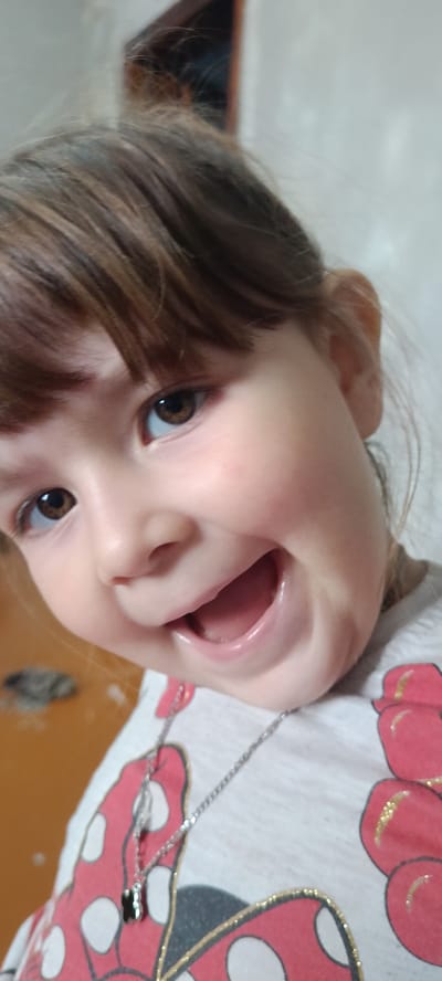 A close-up, eye-level, indoor shot focuses on the face of a young girl in Хохлы, Russia. The girl, with brown hair and brown eyes, is smiling widely, her mouth open. She is wearing a light-colored t-shirt with a Minnie Mouse design. A silver necklace with a small black pendant hangs around her neck. The background is blurred, showing what appears to be a wall and a doorway.