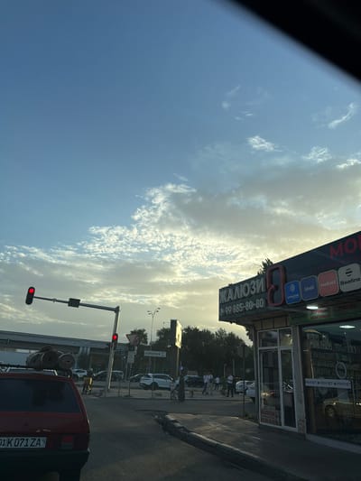 The image depicts a street scene in Uchchinar, Uzbekistan, likely during daylight hours given the clear sky with scattered clouds. In the foreground, the rear of a red automobile is visible, with rolls of carpet secured to its roof. The vehicle's license plate reads "01 K071 ZA".

The scene includes a traffic intersection with a traffic light showing red. A street sign indicates "KORZINKA" with an arrow pointing to the right. Adjacent to the intersection is a storefront displaying the word "ЖАЛЮЗИ" (Jaluzi), meaning blinds, in large lettering. Below this, a phone number "99 885-80-80" is visible. The storefront also features logos for various mobile carriers such as Ucell, mobiuz, and Beeline, suggesting it sells mobile-related accessories.

Several people are visible in the background, walking on the sidewalk and crossing the street. A delivery person on a motorcycle is also present. The environment is urban, with trees lining the street and buildings in the distance. The overall impression is of a typical day in a city, with traffic, commerce, and pedestrian activity.