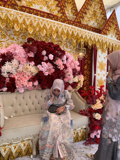 The image depicts an indoor scene, likely a wedding or formal event, characterized by opulent decorations. A woman wearing a hijab and a patterned dress is seated on a tufted white sofa, talking on her phone. She has henna designs on her hands. To her right, another woman, also wearing a hijab and a floral-patterned dress, stands and appears to be looking away from the camera. The background is elaborately decorated with large floral arrangements in shades of pink and red, and ornate gold and red fabric draping. There's also a crystal chandelier visible. The sofa is detailed with diamond-like studs. The floor appears to be marble. The overall ambiance suggests a festive and luxurious setting. The provided image context does not include information about the city of Blukateubai, Indonesia.