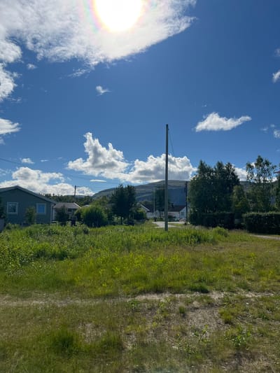 This image depicts a serene, sun-drenched outdoor scene in Lakselv, Norway. The time of day appears to be midday, with the sun casting a bright glow and creating a halo effect on the left side of the frame. The weather is clear and sunny, with a scattering of fluffy white clouds against a vibrant blue sky.

The scene is divided between a lush, green grassy field on the right and a view of scattered houses nestled amongst trees and hills in the distance on the left. A prominent wooden utility pole stands tall in the mid-ground, supporting power lines that stretch across the landscape. Two houses are partially visible in the foreground and mid-ground, with light-colored exteriors and visible windows. The surrounding vegetation is predominantly green and appears to be well-maintained.

In the background, rolling hills or mountains can be seen, partially obscured by trees and the distance. The overall impression is one of a peaceful rural or suburban environment. There is no visible text in the image. The image conveys a sense of calm and pleasant weather, typical of a summer day in a northern climate.