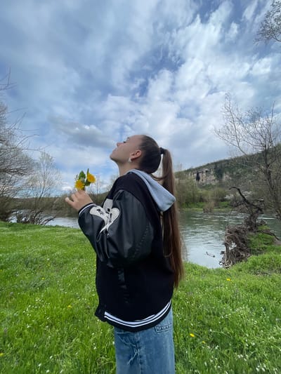 A young woman stands in a grassy area by a river. She has long brown hair pulled back in a ponytail and is wearing jeans and a black jacket with white stripes on the sleeves. She is holding a small bunch of yellow dandelions. The river flows in the background, with a cliff visible on the opposite bank. The sky is cloudy with patches of blue. The scene is the city of Barkach, Bulgaria.