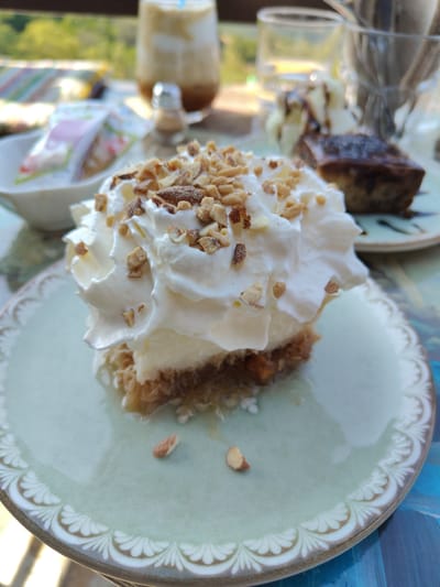 A close-up, eye-level shot features a dessert with whipped cream and chopped nuts, served on a light green plate with an ornate rim. The dessert is layered with what appears to be ice cream or a similar creamy filling over a textured base, possibly pastry or cake. In the background, slightly out of focus, are various items on a table: a glass with a beverage and straw, a salt and pepper shaker, and a piece of chocolate cake. The scene is set outdoors, with blurred green foliage and sunlight indicating a natural environment. The backdrop suggests a cafe or restaurant setting in Palaios Panteleimonas, Greece, a village known for its traditional architecture and scenic views.