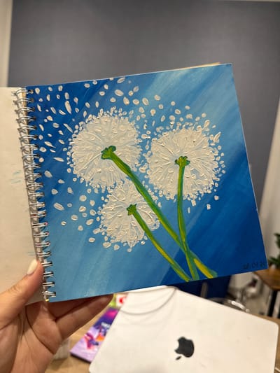 A person's hand, with a light-colored fingernail, holds an open spiral-bound sketchbook. The sketchbook displays a vibrant acrylic painting of three dandelion seed heads against a bright blue background with white flecks that suggest wind-blown seeds. The dandelions are depicted with textured white petals and green stems. In the bottom right corner of the painting, a date "28.01.24" is written in black ink. The background environment is blurred, showing a dark wall, a wooden table, and the corner of a white laptop with an Apple logo. There is no information in the image to suggest it is located in Murom, Russia.