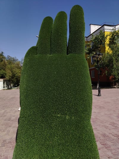 A topiary sculpture shaped like a giant hand with three raised fingers is the central focus of this image, situated in an urban public space in Baikonur, Kazakhstan. The sculpture is entirely covered in dark green, artificial turf, giving it a uniform and manicured appearance. The hand sculpture is positioned on a paved area made of reddish-brown interlocking pavers, arranged in a repeating pattern.
Behind the sculpture, a clear blue sky dominates the upper portion of the frame. To the right, a multi-story building with a bright yellow facade and a red lower section stands. White decorative molding is visible along the roofline of the building. A black, ornate street lamp with two globes is located in front of the building, casting a shadow on the paved ground. Trees with dense green foliage are scattered throughout the background, some appearing on the grounds of the building and others in the wider vicinity. A white, open-air structure, possibly a gazebo or pavilion, is visible on the left side of the image, with trees and a power line in the distance. A person's lower legs and white sneakers are partially visible near the bottom left of the frame, standing beside the hand sculpture. The overall scene depicts a well-lit, outdoor public area on a sunny day.
