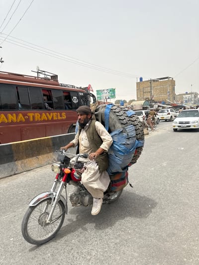 A man wearing a turban, vest, and traditional clothing rides a red motorcycle loaded with a large tire wrapped in blue tarp and a striped blanket. He is in motion on a paved road, appearing to be in a city. In the background, a brown bus with "ANA-TRAVELS" written on its side is visible, along with other vehicles and buildings. The environment suggests a warm, possibly dusty day with a bright sky. The scene is set in Kuchlak, Pakistan.