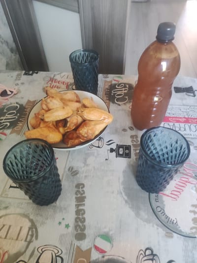 A plate of chicken wings, a large plastic bottle of a brown beverage, and three dark blue textured glasses are arranged on a table covered with a patterned tablecloth. One glass is near the top right of the image, another is to the left of the chicken wings, and the third is in the bottom right corner. The chicken wings are golden brown and appear to be cooked. The bottle is a standard plastic beverage bottle with a dark cap. The tablecloth has various printed designs, including text that appears to be in Italian, coffee-related imagery, and what looks like the Italian flag. The background shows a wooden cabinet or wall. The scene is indoors and appears to be a casual meal setting. The image does not contain any information that would allow for the determination of its location within the city of Kovel, Ukraine.