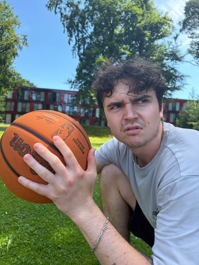 A young man with curly brown hair and a slightly furrowed brow kneels on a grassy lawn, holding an orange basketball. He is wearing a light grey t-shirt and dark shorts, and a silver bracelet adorns his left wrist. The man is looking off to the right of the frame with a serious expression. In the background, a modern building with red facade and large windows stands amongst lush green trees under a clear blue sky. The scene appears to be an outdoor recreational area, possibly a park or a sports field. The location is in the city of Enschede, Netherlands.