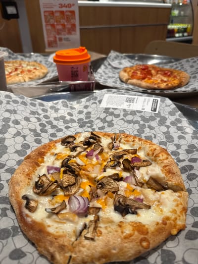 A close-up, eye-level shot features a mushroom and red onion pizza with melted cheese, served on a decorative paper liner. In the background, a small pizza with tomatoes and cheese is visible, along with an orange-lidded cup and a poster displaying prices and images of pizzas, suggesting a fast-food restaurant or pizzeria. The scene appears to be indoors, possibly in a casual dining establishment in the city of Chusovoy, Russia, given the context. The lighting is dim, with a focus on the food.