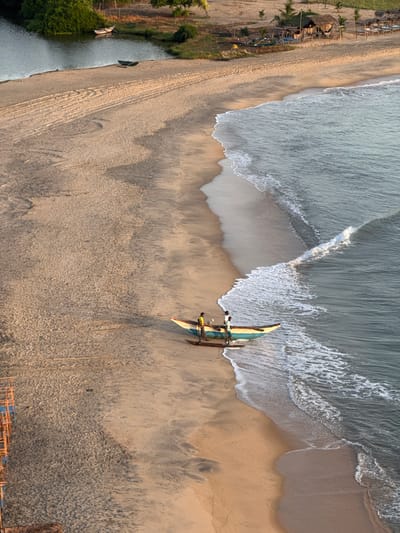 This is an aerial, high-angle shot of a beach scene in Shastrawela, Sri Lanka, taken during what appears to be either sunrise or sunset given the warm, soft light casting long shadows. The image is oriented vertically, with the ocean on the right and a beach separating it from a river and vegetation on the left.

The ocean features gentle waves with white foam washing onto the sandy shore. On the beach, a traditional outrigger canoe, painted in teal and white with red accents, is positioned near the water's edge. Three people, dressed in light-colored clothing, are visible in or near the canoe, suggesting they are either preparing to launch, have just returned, or are resting after a fishing trip. Their activity is not definitively clear from this still image.

Further left, a narrow river or inlet runs parallel to the beach, with at least two smaller boats moored along its bank. Dense green vegetation lines the river, contrasting with the dry sand of the beach. Along the edge of the beach, near the vegetation, are several simple wooden structures with thatched roofs, possibly huts or temporary shelters. Tire tracks or footprints are visible in the sand, indicating human activity.

The overall mood is peaceful and serene, capturing a moment of coastal life in Sri Lanka. There is no visible text in the image.