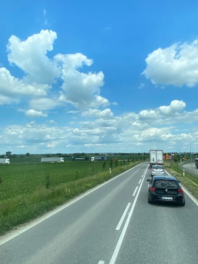 In Podłęże, Poland, on a sunny day with a bright blue sky dotted with fluffy white clouds, a traffic jam is visible on a highway. The road features white lane markings. In the foreground, a black BMW hatchback with license plate "KWI 3112H" is stationary, followed by a silver hatchback and a white truck. Beyond the road, a vast green field stretches towards the horizon, with several white and green trucks and trailers parked along the roadside in the distance. Trees are scattered throughout the landscape, and the overall scene suggests a rural or semi-rural setting.