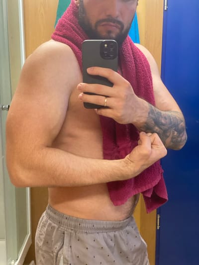 A man is standing in front of a mirror, taking a selfie. He is shirtless and wearing gray shorts with a pattern. A pink towel is draped around his neck. His right arm is flexed, showing his bicep. He has a beard and is wearing a ring on his left hand. There is a tattoo on his left forearm. The background appears to be a locker room, with a blue door and wooden walls. The lighting is artificial. The image is taken from the chest up.