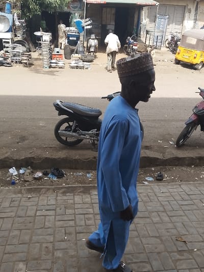 A dark-skinned man, wearing a light blue traditional tunic and a patterned cap, walks right to left on a paved sidewalk in Kofar Mazugal, Nigeria. His mouth is slightly open, and his gaze is directed forward. Behind him, a black motorcycle is parked on an unpaved dirt shoulder littered with debris, which separates the sidewalk from an asphalt road. Another red motorcycle and a yellow motorized tricycle are also visible on the street. Across the road, a row of commercial buildings features a shop displaying numerous metal cooking pots, basins, and other items outside. A "PEPSI" sign and visible phone numbers ("0701817766", "0701124784") are mounted above the shopfronts. Two men are observable near the first shop: one stands near the entrance, and another walks away from the viewer further down the street. The environment appears to be a busy commercial area.