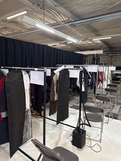 The image depicts an indoor scene within an industrial-style building in Herentals, Belgium, likely a warehouse or event space. The primary focus is an arrangement of multiple metal garment racks positioned in the foreground and mid-ground. These racks are laden with various clothing items, many enclosed in black garment bags or clear plastic. White rectangular labels are affixed to the racks.

In the immediate foreground, a dark gray folding chair is partially visible on the bottom left. Another similar folding chair is situated in the mid-right, adjacent to a black garment steamer unit with its hose and nozzle extending upwards. A power cord from the steamer extends across the light-colored floor. A row of additional dark gray folding chairs is visible further back, behind the main clothing racks.

A continuous black curtain serves as a backdrop, spanning the mid-ground horizontally. Above the curtain, the high industrial ceiling is visible, featuring exposed metal trusses, corrugated metal panels, and utility installations including HVAC ducts, electrical conduits, and multiple suspended, rectangular fluorescent or LED light fixtures. In the far background, to the right of the black curtain, a partial view of a black Mercedes-Benz commercial vehicle is discernible, along with additional industrial equipment. No individuals are present or identifiable within the frame. The scene suggests a temporary setup, possibly for a fashion event, photoshoot, or wardrobe preparation.