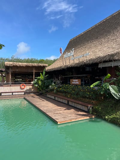 The image depicts a serene waterside cafe with a thatched roof, located in Tambon Pa Kae Bo Hin, Thailand. The cafe features a rustic design with bamboo accents and a wooden deck extending over turquoise water. Lush greenery surrounds the establishment, with a variety of plants and flowers adding to the tropical ambiance. A life preserver is visible hanging on a bamboo post, suggesting proximity to the water. In the background, a dense forest creates a natural backdrop. The sky is a vibrant blue with scattered white clouds, indicating a clear day. A sign with Thai script is displayed on the cafe, likely indicating its name or services. The overall scene conveys a tranquil and inviting atmosphere.