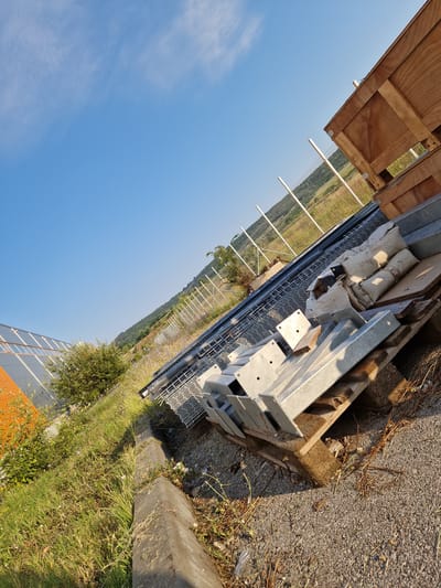 The image depicts an outdoor scene, likely a construction or storage area, on a sunny day. In the foreground, a pallet laden with metal components, possibly for solar panel installation or construction, is visible. The metal pieces are primarily silver and appear to be brackets and supports. Some of the components are covered with white protective material. To the left of the metal components, a gray concrete curb lines a grassy embankment, with a modern building with orange and gray paneling in the background. Beyond the building, a dense green bush adds to the vegetation. A wire mesh fence runs diagonally across the mid-ground, separating the foreground from a hillside covered in trees and vegetation in the background. The sky is a clear blue with sparse white clouds. The overall impression is one of an unfinished project or a staging area for materials. The location is identified as Bahovitsa, Bulgaria.