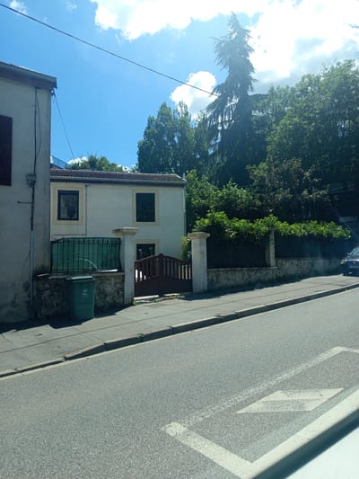 The image depicts a street scene in Nancy, France, on a sunny day. The perspective is from a slightly elevated position, looking down towards the street and the buildings lining it. The sky is bright blue with some scattered white clouds, suggesting it is daytime, likely late morning or early afternoon.
In the foreground, the asphalt of the street is visible, with white road markings, including a diamond shape, painted on its surface. A sidewalk runs alongside the street, bordered by a low wall. To the left of the sidewalk, a green trash bin is placed against a stone wall.
Adjacent to the sidewalk and wall is a two-story building with a light-colored facade. It has two windows on the upper floor and two on the lower floor, all with dark frames. A green wrought-iron gate is visible on the lower part of the building, and above it, a wooden bench is situated on a small porch or balcony. Trees and foliage are abundant, with dense green leaves from a large tree dominating the left side of the frame, creating a sense of a residential or suburban area. The roof of another building, with reddish tiles, is partially visible in the lower right corner. The overall impression is of a quiet, pleasant day in a European town. There is no visible text in the image.