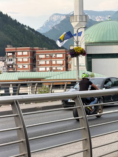A vertical shot depicts a scene in Konjic, Bosnia and Herzegovina. In the foreground, a modern metal railing runs along the right side of the image. Beyond the railing, a person on a motorcycle, wearing a black t-shirt and a green and white helmet, is visible. The motorcycle itself appears to be a dark color.
Behind the motorcycle, a dark-colored car is parked, its windows tinted. To the left of the motorcycle, a white minaret stands tall, topped with a conical structure. Several flags, including one with yellow stars on a blue background and another with blue and white stripes, are attached to a pole extending from the minaret. A basket filled with colorful flowers, featuring yellow and purple blooms, hangs near the flags.
Further in the background, a large, pale green dome dominates the upper left portion of the image, suggesting a significant building such as a mosque or cathedral. Adjacent to the dome are multi-story brick buildings with numerous windows, some of which have air conditioning units visible. The landscape beyond the buildings shows a densely forested mountainside with a hazy sky. The overall impression is that of an urban environment nestled within a natural, mountainous setting. The lighting suggests it is daytime, likely overcast due to the soft light and lack of strong shadows.