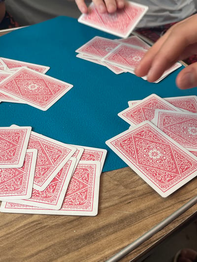 The scene depicts an indoor card game in progress in Barabinsk, Russia. A significant number of red-backed playing cards, each adorned with a white intricate floral and geometric pattern, are distributed across a circular teal-blue playing mat. This mat partially covers a light brown wooden table featuring a visible grain. At least three individuals are present. One person, wearing a light-colored top, has both hands visible at the top-center of the frame, holding a single card mid-air above a stack of face-down cards. Another individual's right hand, with fingers slightly spread and blurred from motion, is positioned over a separate cluster of cards on the right side of the mat; a patterned garment is visible behind this hand. A third person's patterned sleeve is partially visible on the far left. Multiple piles and scattered cards are distributed across the mat, indicating active gameplay. A thin, silver-colored metallic rod or rail is situated on the lower right edge of the wooden table.