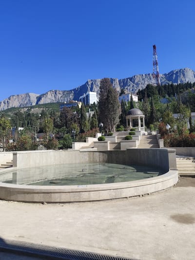 The image depicts a large, circular, shallow fountain in an outdoor setting. The fountain is made of light-colored stone and contains clear water. Behind the fountain are multiple tiers of stone steps leading up to a small gazebo-like structure with a domed roof, also made of light-colored stone. Various trees and shrubs are scattered throughout the scene, and buildings are visible in the mid-ground. In the background, a tall mountain range is present, with a red and white communications tower on one of the peaks. The sky is clear and blue. The scene appears to be located in a park or garden area. There is no evidence to suggest the location is in George, South Africa. The scene is serene and well-maintained. No people are visible in the immediate foreground or mid-ground.
