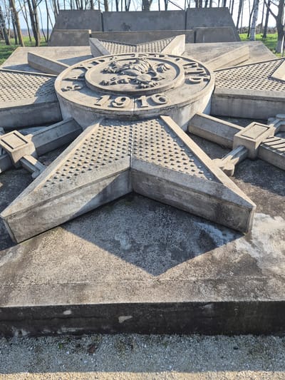 The image shows a stone monument in Shumentsi, Bulgaria. The monument is a large, stylized Maltese cross with a circular medallion in the center. The medallion features a relief image and the inscription "1916." The cross has a textured surface with small, rounded protrusions. The monument is made of grey stone and is partially in shadow. The background includes trees and a gravel surface.