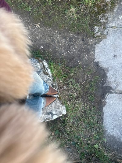 The image displays a top-down view of a person standing on a large, irregular light-gray stone block in Cernay-la-Ville, France. The individual wears light blue denim jeans and brown leather boots with pointed toes. A blurred, light-tan, fibrous material occupies the immediate foreground, obscuring the upper part of the individual and scene. The stone block, stained with dark lichen or moss, is situated on ground composed of patchy green grass, exposed dark soil, and scattered small debris. To the left of the stone and person's feet, a narrow path of dark, compacted gravel or dirt is visible. To the right of the grassy area, a section of light gray concrete paving stones forms another distinct surface. The lighting suggests natural daylight.