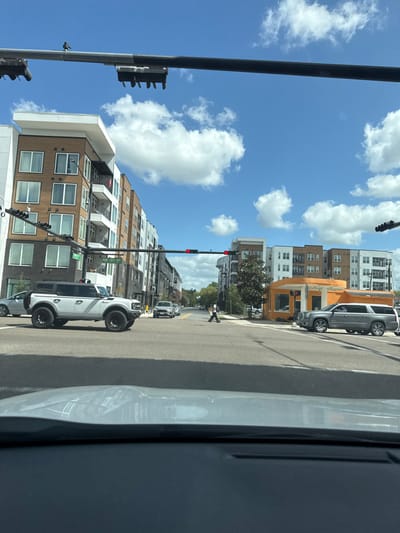 The image captures an urban intersection in Gainesville, Florida, USA, specifically the junction of NW 1st Street and NW 2nd Avenue. The perspective is from inside a stationary vehicle, looking generally eastward under a bright blue sky with scattered white cumulus clouds.

In the immediate foreground, the top of a vehicle's dashboard and windshield is visible. Overhead, a black gantry supports multiple traffic light fixtures, displaying red signals for the observer's direction. A small, dark bird is perched on the upper-left section of this gantry.

At the intersection, a white four-door Ford Bronco, notably lifted with dark aftermarket wheels, is positioned in the left lane, slightly past the stop line, oriented away from the camera. In the right lane of the perpendicular street, a long, grey SUV, identified as a GMC Yukon XL or similar extended vehicle, is stopped at the crosswalk. A single pedestrian, dressed in dark clothing, is actively crossing the street from right to left, positioned between the white Bronco and the grey SUV.

To the left of the intersection stands a modern multi-story apartment building featuring a facade of white and light brown paneling, with numerous windows and balconies. On the right corner of the intersection, a distinctive, small, single-story orange-colored commercial building, possibly a repurposed structure such as a former gas station, is visible. Behind it and extending down the street are additional multi-story residential or mixed-use buildings with various contemporary architectural styles. A green street sign indicating "NW 2ND AVE" and "NW 1ST ST" is mounted on a pole near the left side of the intersection.