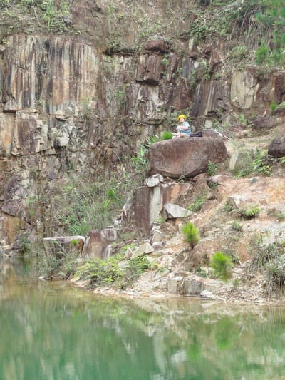 The image depicts a person wearing a yellow hard hat sitting on a large rock near a body of water. The environment appears to be a quarry or a natural rock formation with a steep, rocky cliff face. Vegetation, including grasses and small trees, grows sparsely on the rocks and along the water's edge. The water is a greenish hue, and its surface reflects the surrounding landscape. The person is facing towards the left of the frame, and a dark bag is placed on the rock next to them. The location is identified as Lán Tranh, Vietnam.