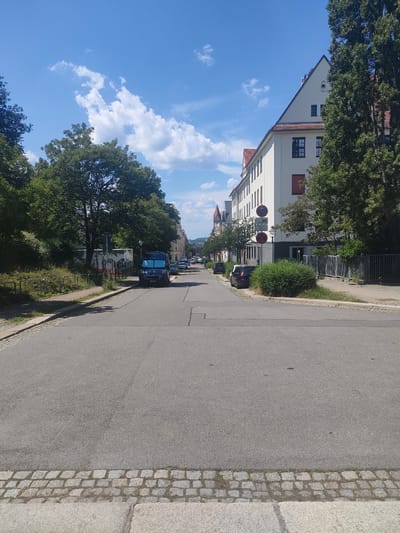 The image shows a street intersection in Chemnitz, Germany, on a sunny day. The foreground is dominated by a paved road with cobblestone edging. To the right, a white building with a red-tiled roof and multiple windows lines the street. Several trees with green foliage are visible on the left side of the road, and a dark blue van is parked on the right. Further down the street, more buildings and trees can be seen, along with other parked cars. The sky is bright blue with scattered white clouds. The scene appears to be a residential or mixed-use area in the city.