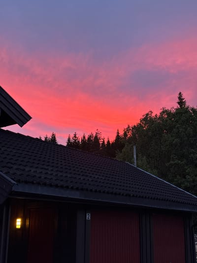 A residential property at Åmot, Norway, is depicted at dusk. The sky is illuminated with vibrant pink and purple hues, indicative of a sunset. In the foreground, a dark-tiled roof of a building, possibly a garage or house, dominates the lower portion of the image. The building features red doors and a yellow outdoor light fixture. A number "2" is visible on a vertical post. Behind the building, a line of coniferous trees forms a silhouette against the colorful sky, with denser foliage of deciduous trees to the right. The overall scene captures the serene beauty of a twilight sky over a dwelling in a natural setting.