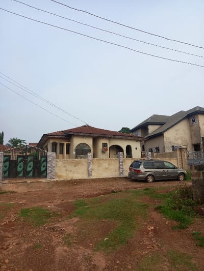 The image shows a residential area in Ita Oniyan, Nigeria, located at latitude 7.293 and longitude 5.169. There is a single-story house with a brown roof and a beige facade. The house has a large, open-air porch with a decorative archway. There is a grey minivan parked in front of the house. The car is facing the street, and there are several power lines running across the sky. The house has a black gate with green panels on the front. The front yard has a patch of grass, and the rest is dirt. There are no people present in the image. The image was taken during the day, and the sky is clear and blue.