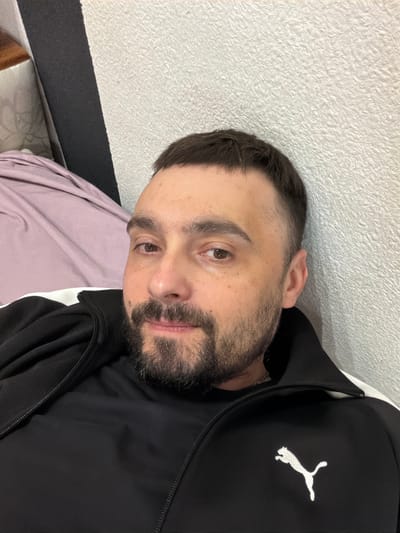A man with short dark hair and a beard is lying on his side on a bed with a light purple sheet. He is wearing a black Puma track jacket with a white logo on the right chest. The background is a white wall. There is no information in the image to suggest a location in the city of Kovel, Ukraine.