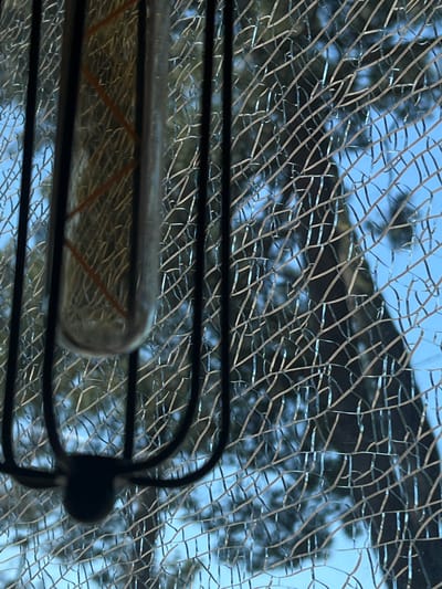 A close-up shot captures a broken window with a spider-like object on the lower left. The glass is severely cracked, creating a web-like pattern. Through the fractured panes, a blurry background of trees and a blue sky is visible. The object appears to be a black metal cage with a glass bulb inside, resembling a light fixture. The overall scene is dark and the lighting is low, with the only light source seemingly coming from the background. The image does not depict any specific location within the city of Ochamchire, Abkhazia.