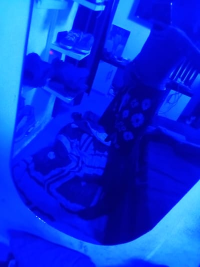 The image is a dimly lit, vertically oriented photograph taken in Ogbogbo, Nigeria, under intense blue lighting. The scene appears to be an indoor setting, possibly a bedroom or storage area. In the foreground, a blue tub or basin is visible, filled with a dark, patterned material. The pattern on the material consists of white floral designs on a dark background.
On a shelf above the tub, a pair of dark-colored sneakers is placed. These sneakers have white laces. Behind the sneakers, more shelves are visible, holding various indistinct objects. To the left of the shelves, a portion of a light-colored wall or partition is present. The overall impression is one of a cluttered, dimly lit space. The blue lighting creates a surreal and somewhat disorienting atmosphere. There are no people visible in the image.