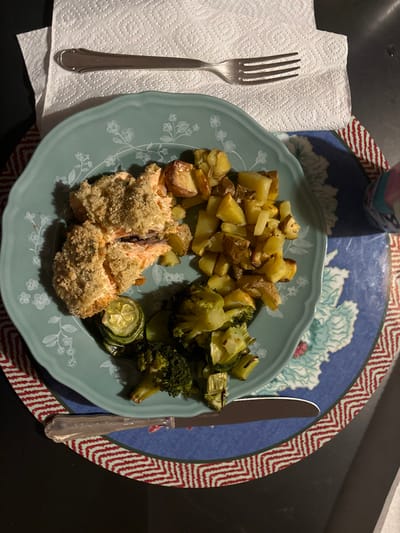 A blue plate with a white floral design sits on a red and white patterned placemat on a black surface. The plate contains a meal consisting of roasted salmon, baked potatoes, broccoli, and zucchini. A silver fork lies on a white paper napkin to the left of the plate. A silver knife is positioned below the plate. The meal is located at 46.514 degrees latitude and 9.859 degrees longitude, in Celerina/Schlarigna, Switzerland.