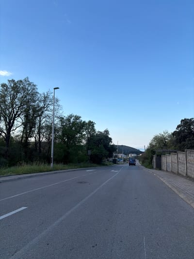 The image depicts an asphalt road in Zgrade, Montenegro, curving uphill to the right. A blue truck is visible further up the road. White lane markings are present on the road surface. Trees and vegetation line the left side of the road, along with a tall street lamp. On the right side of the road is a concrete barrier and more vegetation. The sky is clear and blue.