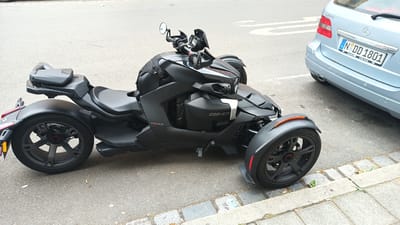 The media file shows a black three-wheeled Can-Am Ryker motorcycle parked on a paved street. The motorcycle is in the foreground, facing slightly to the right. It has a matte black finish and features a prominent front end with distinctive headlights and a windshield. The handlebars are visible, along with mirrors and a small digital display. The vehicle has two wheels at the front and one at the rear. The rear wheel is large with a dark gray, multi-spoke rim and a brake disc with a red accent. A black seat is positioned behind the handlebars, and a small storage compartment is mounted behind the seat. The "Can-Am" logo is visible on the side of the motorcycle, as is the "Ryker" branding.
To the right of the motorcycle, the rear of a light blue Mercedes-Benz B-Class car is visible. The car has a German license plate with the text "N DD 1801". The car is also parked on the street, parallel to the motorcycle.
The scene appears to be outdoors on a paved street with a sidewalk made of gray bricks and stones to the right. There are small patches of grass growing through the cracks in the sidewalk. The asphalt of the street shows some wear and tear, with a few small pieces of debris scattered on it.
The lighting suggests it is daytime, likely under overcast or partly cloudy skies, as there are no strong shadows. The overall mood of the image is neutral, documenting the parked vehicles in a typical urban or suburban street environment. The location context provided is Nuremberg, Germany.