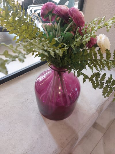 A close-up shot captures a bouquet of artificial flowers in a vibrant magenta glass vase. The flowers, a mix of deep pink and a single cream-colored bloom, are interspersed with delicate green foliage, resembling fern leaves. The vase is positioned on a textured, light-colored surface, possibly a windowsill or shelf, with a dark horizontal element visible behind it. The background is blurred, suggesting an indoor setting with natural light filtering in. The specific location in Kyzylorda, Kazakhstan, is not identifiable from the visual information provided.