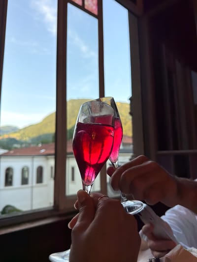 Two champagne flutes filled with a vibrant red liquid are being held up in a toast, their contents catching the light. In the background, through a large window, a scenic view of San Pellegrino Terme, Italy, is visible. The window frames rolling green hills dotted with buildings, and a bright blue sky with sparse clouds. The overall impression is one of celebration and enjoyment, possibly at a restaurant or a similar establishment in the Italian town.