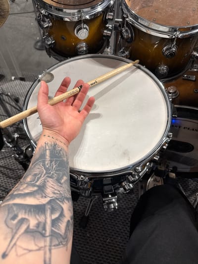 The image shows a section of a drum set, including at least three drums. One drum, centrally located, has a white drumhead and chrome hardware. Adjacent to it are drums with a wood-toned finish and chrome hardware. A hand holds a drumstick near the center drum. The arm connected to the hand has a detailed black and grey tattoo. The background includes carpet and parts of additional drums. There is no information to determine any location.