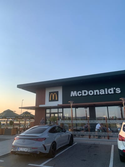 The image shows the exterior of a McDonald's restaurant in Ub Municipality, Serbia. A modern-looking building with large windows and a dark-colored overhang featuring the "McDonald's" logo is visible. The Golden Arches logo is also present on the building.
In the foreground, a sleek, grey Mercedes-AMG car is parked in a designated spot, with its rear facing the camera. The car has a license plate that reads "NS 900-MS". Through the restaurant's windows, people can be seen sitting at tables, suggesting that customers are dining inside. The scene appears to be during the late afternoon or early evening, as indicated by the warm, soft lighting and the clear sky. A few McDonald's umbrellas are also visible outside the restaurant. The overall impression is of a typical fast-food dining experience in a suburban or town setting.