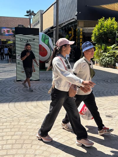In Ashdod, Israel, on a sunny day, two individuals walk across a paved plaza bathed in dappled sunlight. In the foreground, a person wearing black pants and light pink sneakers walks from right to left, facing away from the camera. To their left and slightly behind, another person in black pants and a light pink cap walks in the same direction, also facing away. Behind them, a person in a blue bucket hat and white shirt walks forward, looking to the right. Further in the background, a man in a black shirt and ripped shorts is standing near a large, illuminated display panel with horizontal white lines and a colorful graphic, holding a phone to his ear. To the right of the display, a large soccer ball with green and red panels is visible. The environment includes modern building facades with signage, green foliage, and sunlight casting shadows on the ground.