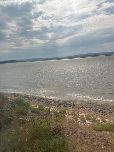 The media file depicts a scenic view of a body of water, likely a lake or a bay, with a shoreline of pebbles and sparse vegetation. The water surface is shimmering with sunlight, suggesting it is daytime. In the distance, a hazy mountain range or landmass is visible, forming a horizon line. The sky above is filled with dynamic clouds, with patches of blue sky interspersed among them, indicating variable weather conditions. The lighting suggests it could be late morning or early afternoon. The scene is tranquil and natural, evoking a sense of peace and openness. The presence of water, land, and sky suggests an outdoor, potentially natural, landscape setting. The image is taken from a slightly elevated perspective, possibly from a car or a hill overlooking the water. There is no visible text in the image.