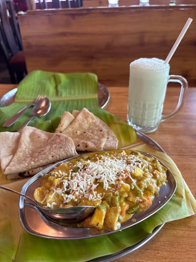 The image captures a meal presentation likely in Kamalapura, India, evidenced by the cuisine and style of serving. On a wooden surface, there's a layered serving arrangement. At the base is a metal tray layered with broad green banana leaves. On the right, a silver oblong dish contains a yellow, creamy vegetable curry topped with shredded white cheese. A spoon is inserted into the curry. To the left of the curry, stacks of chapati bread are neatly placed on the banana leaves. Beside the chapati, a spoon and a fork rest on the green leaves. In the background is a glass mug filled with a frothy white beverage, possibly lassi, and a white straw. The mug has a handle and sits atop the edge of the tray. The light suggests daytime. The environment appears to be a restaurant or eatery due to the presentation style. No people are visible in the image.