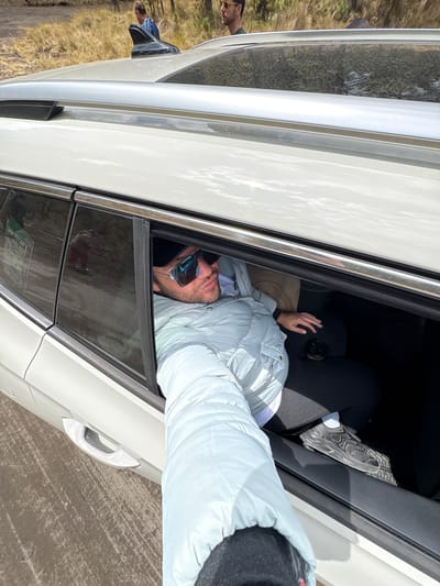 A man with a beard and mustache, wearing a white jacket and gray beanie, is hanging out of the open window of a white car. He is holding a cell phone to his ear. Inside the car, another person wearing black pants and gray sneakers can be seen. The car appears to be parked on a paved surface. In the background, there is a grassy area and a person walking. The license plate indicates that the location is near Popocatépetl, Mexico.