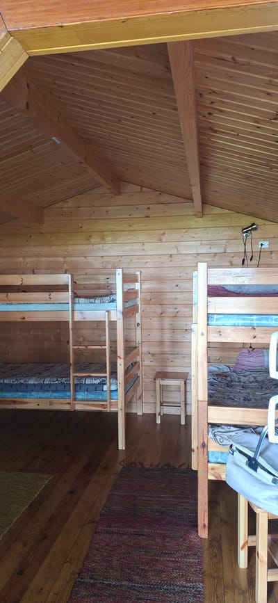 The image depicts the interior of a wooden cabin or room with bunk beds. The walls and ceiling are constructed from horizontal wooden planks, and the ceiling has exposed wooden beams. Two sets of wooden bunk beds are visible. The bed on the left has mattresses covered in blue and patterned fabric. The bed on the right has mattresses with blue and patterned fabric, and one mattress is topped with a purple and white patterned pillow. A small wooden stool is positioned between the two sets of bunk beds. A rug with a red and dark blue patterned weave lies on the wooden floor in the foreground. Another rug with a yellow-green weave is partially visible in the lower left corner. A floor lamp with a metal shade is mounted on the wall above the right set of bunk beds, and an electrical outlet is also visible. In the bottom right corner, part of a grey and white bag or carrier is resting on the floor near the legs of the right bunk bed. The scene appears to be a lodging facility, possibly a campsite or hostel, located in Valma, Estonia. There are no people visible in the image, and no specific actions or interactions are depicted.