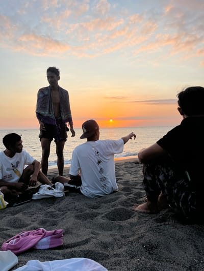 The image displays four male individuals on a dark sand beach in Desa Batu Layar, Indonesia, during sunset. The sun, a bright orange disc, is low on the western horizon over the calm ocean, creating an orange and pink luminescence across the sky and reflecting on the water. The sky above features scattered, thin clouds.
From left to right, the individuals are positioned as follows:
1. A male is seated on the dark sand, dressed in a white t-shirt with a graphic depicting a bird and the text "SOUTH DAKOTA," and dark shorts. He faces partially right.
2. A male stands barefoot at the water's edge, shirtless, wearing dark shorts, with a multi-colored, patterned sarong draped over his shoulders. He faces partially left.
3. A male is seated on the sand with his back partially to the viewer, wearing a white t-shirt with black abstract symbols on the back and a dark baseball cap worn backward. He extends his right arm, pointing directly towards the setting sun over the ocean.
4. A male is seated on the sand with his back to the viewer, dressed in a black t-shirt and dark, patterned pants. His bare feet are visible in the sand.
Miscellaneous clothing items, including a pink shirt and a white shirt/towel, are visible on the sand in the foreground. The group is observing the sunset over the ocean.