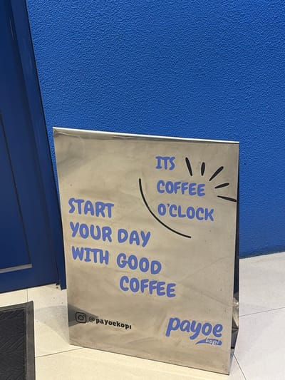 A metal sign is propped up against a textured blue wall. The sign displays text and illustrations related to coffee. At the top, there are stylized lines resembling a sun or rays, with the words "ITS COFFEE O'CLOCK" written below in blue, bubbly lettering. To the right, the word "payoe" is written vertically in larger blue letters, with "kopi" written below it in smaller print. At the bottom of the sign, the words "START YOUR DAY WITH GOOD COFFEE" are written in blue letters. There is also an Instagram logo and the handle "@payoekopi" on the right side of the sign. The lighting suggests it might be daytime. The scene appears to be an outdoor or semi-outdoor area, possibly a cafe or shop entrance, given the presence of a blue door or wall and floor tiling. The overall impression is a cheerful and inviting message promoting coffee.